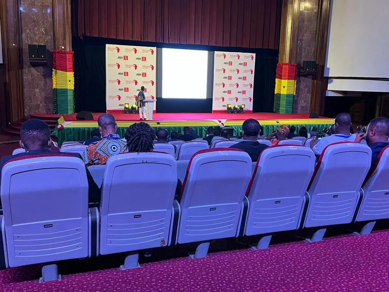 A speaker addressing delegates from the stage at a SASCON plenary in Accra, between conference backdrops and Ghana flag drapes.