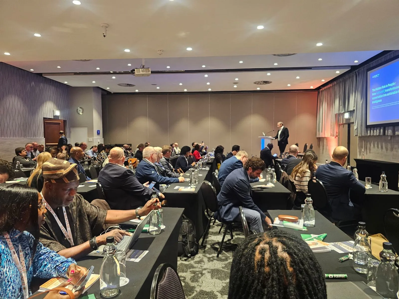 Delegates seated at long tables in a SASCON session, working with laptops, notebooks and printed papers while a speaker presents from a lectern beside a projected slide.