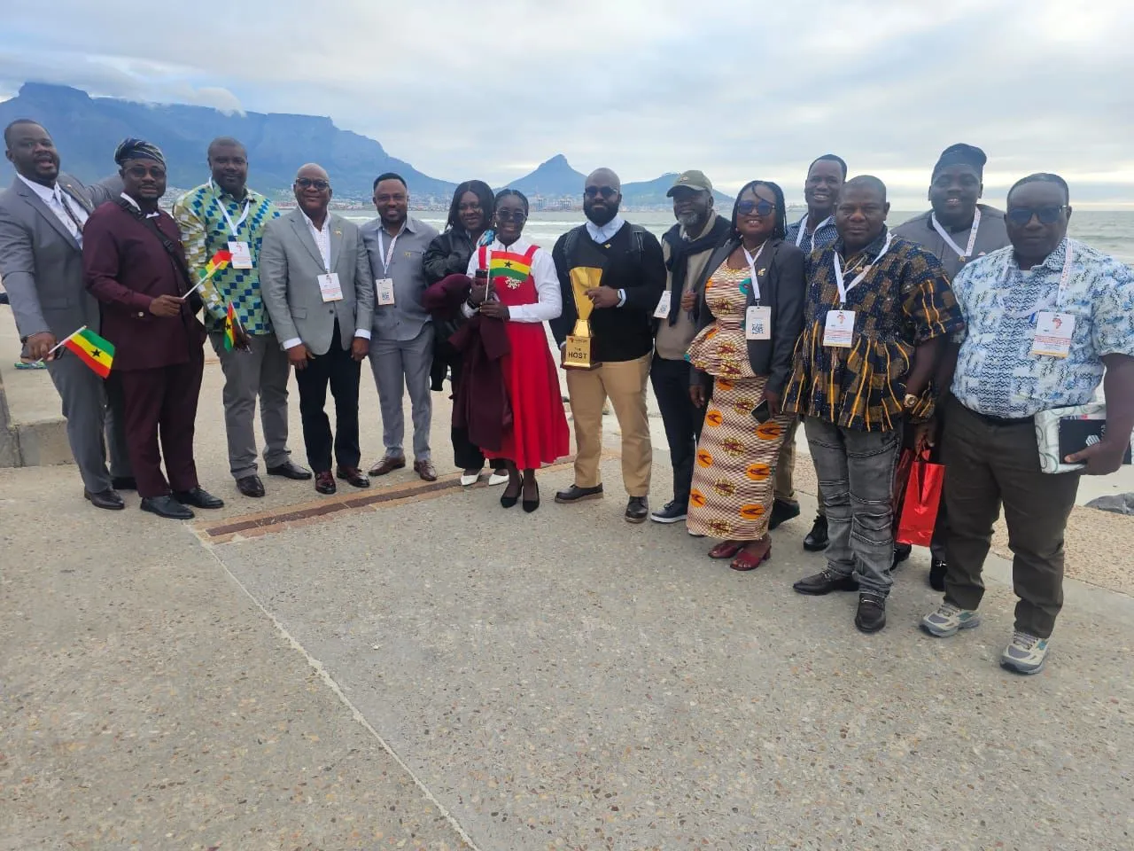 The Ghanaian delegation at the Cape Town edition of SASCON, standing together on the waterfront with Table Mountain behind them, holding the host trophy and small Ghana flags.